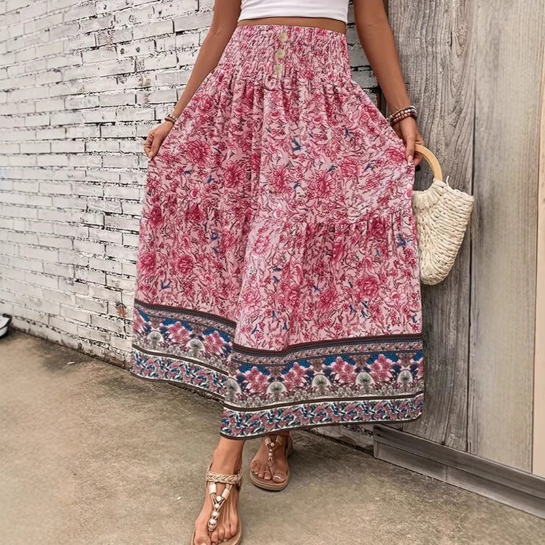 Femme portant une jupe longue fleurie rose avec motifs floraux, sandales beiges, sac en paille, posant devant un mur en briques blanches.