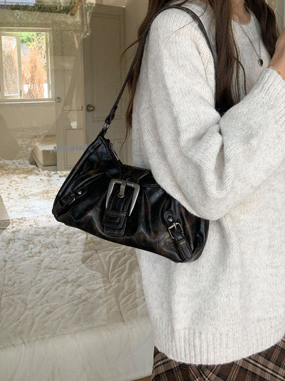 Femme portant un pull beige et un sac à main noir en cuir avec boucle argentée, reflet dans une vitre, intérieur lumineux en arrière-plan.