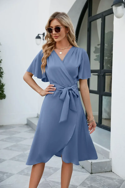 Femme en robe bleue élégante, manches courtes, ceinture nouée, lunettes de soleil, posant devant une porte moderne. Mode féminine chic, style estival.