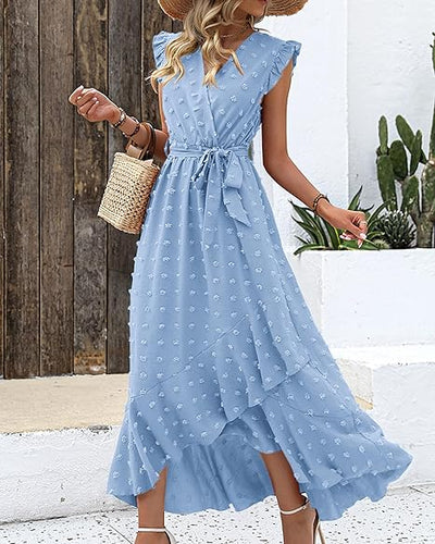 Robe longue bleue à volants, motif floral, portée par une femme avec chapeau de paille et sac en osier, devant un mur en bois et plantes vertes.