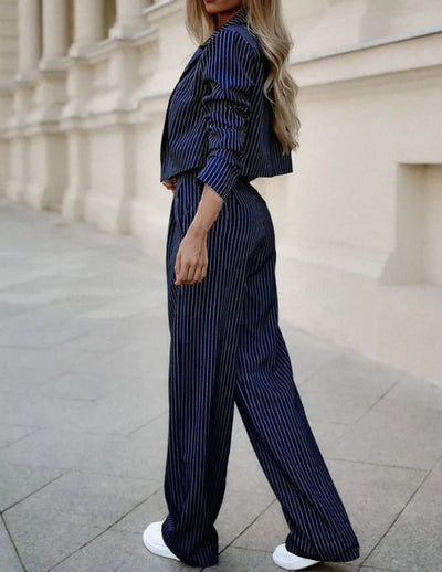 Femme en tailleur pantalon rayé bleu marine, cheveux longs blonds, pose élégante sur fond urbain, chaussures blanches, mode chic et moderne.