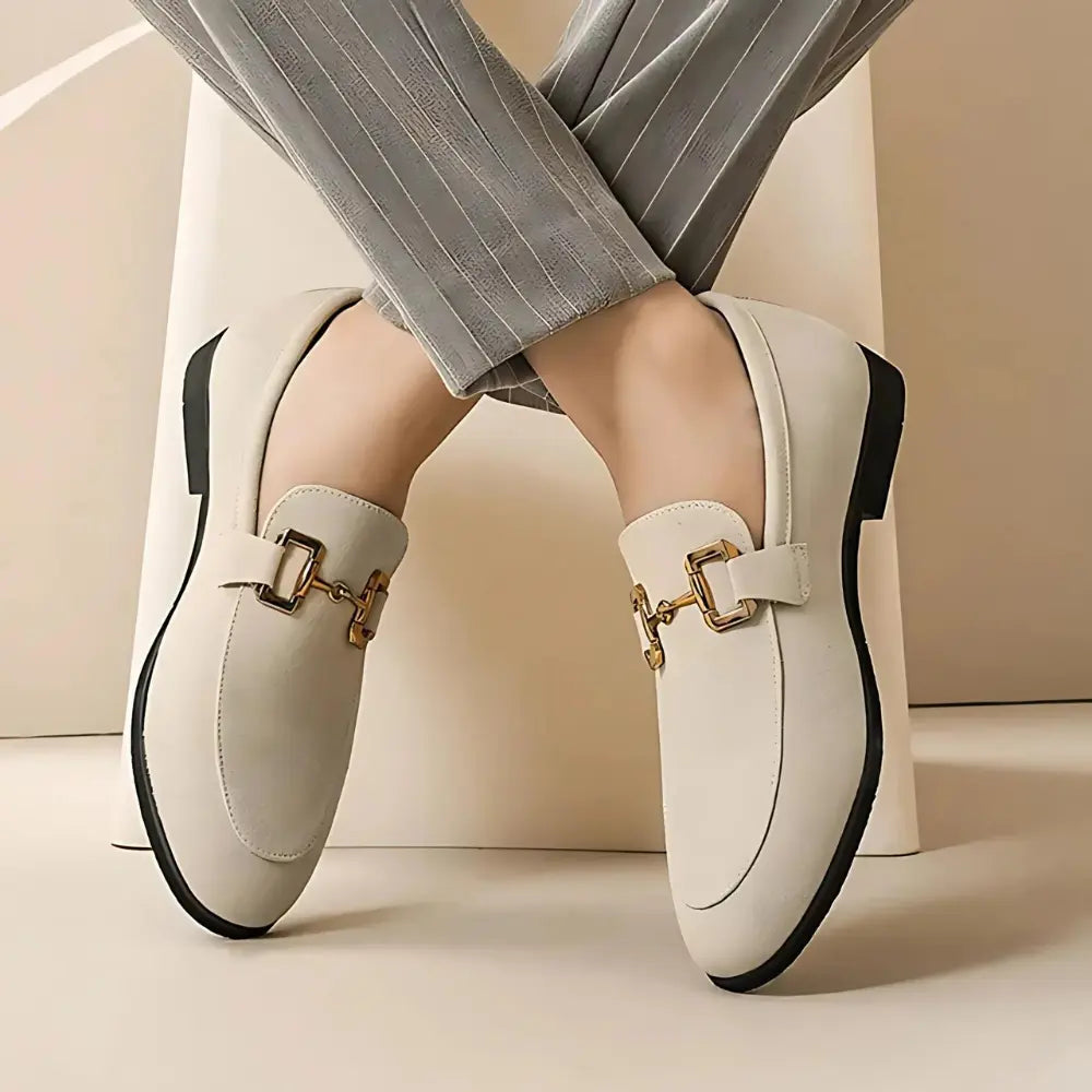 Chaussures mocassins beiges élégantes pour femmes avec boucle dorée, portées avec un pantalon à rayures grises. Mode féminine chic et moderne.