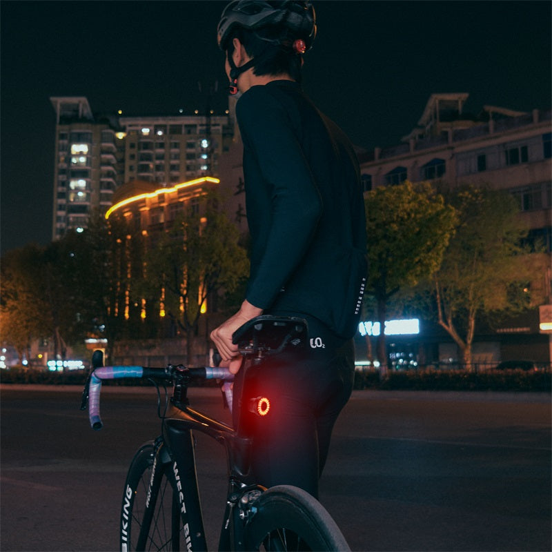Cycliste de nuit en tenue noire avec casque, tenant un vélo de route équipé d'une lumière arrière rouge, en ville illuminée. Sécurité cycliste nocturne.