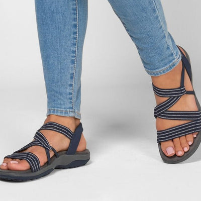 Jeans bleus et sandales de randonnée noires avec lanières croisées, portées par une personne debout sur un sol blanc. Mode décontractée et confortable.