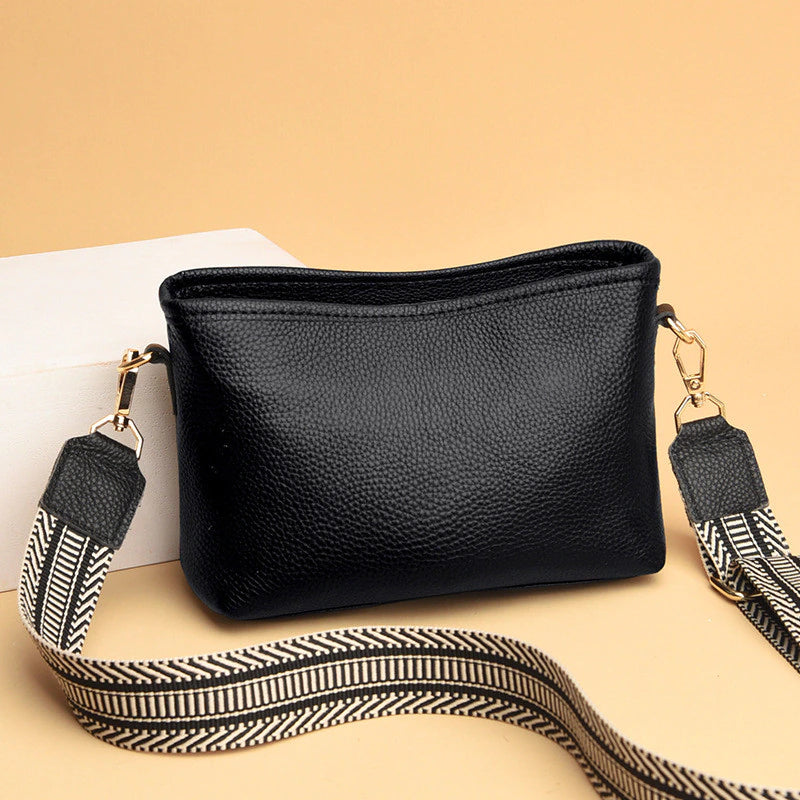 Sac à bandoulière noir en cuir texturé avec sangle ajustable à motifs géométriques, posé sur fond beige. Accessoire mode élégant et pratique.