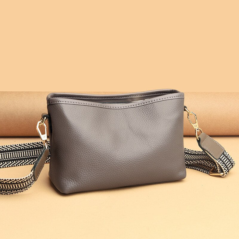 Sac à bandoulière en cuir gris avec sangle ajustable à motif chevron, posé sur fond beige. Accessoire mode élégant et pratique pour femme.