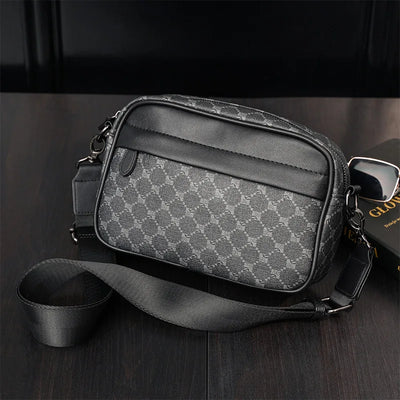 Sacoche en cuir noir et gris avec motif géométrique, bandoulière ajustable, posée sur une table en bois sombre. Accessoire mode homme élégant.