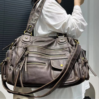 Sac à main en cuir marron avec multiples poches zippées, porté par une personne en chemise blanche. Accessoire mode élégant et pratique.