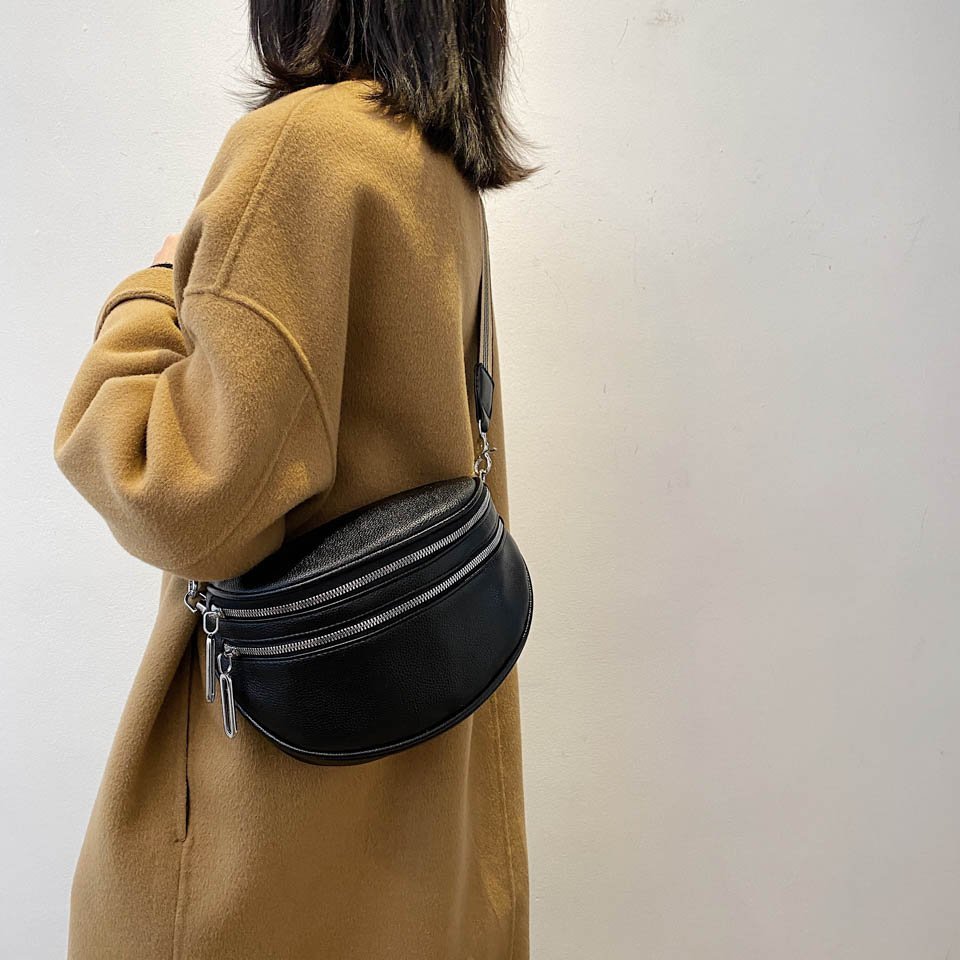 Femme portant un manteau camel et un sac banane noir en cuir avec fermetures éclair argentées, mode automne-hiver, style urbain chic.