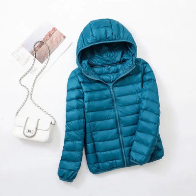 Veste doudoune bleue avec capuche sur fond blanc, accompagnée d'un sac à main blanc à chaîne argentée et d'un magazine. Mode hiver tendance.