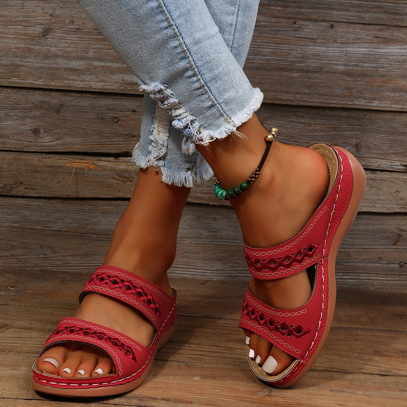 Sandales rouges en cuir pour femme avec lanières ajourées, portées avec un jean déchiré et un bracelet de cheville, posées sur un fond en bois.