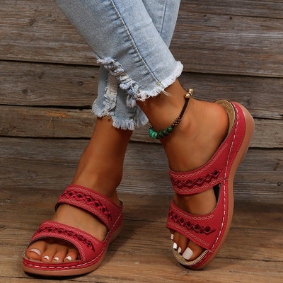 Sandales rouges en cuir pour femme avec lanières ajourées, portées avec un jean déchiré et un bracelet de cheville, posées sur un fond en bois.
