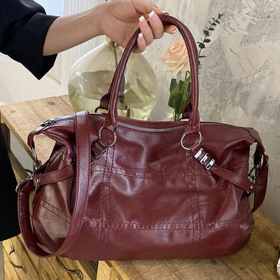 Sac à main en cuir rouge bordeaux, design élégant avec poignées doubles et bandoulière, posé sur une table en bois, décor floral en arrière-plan.