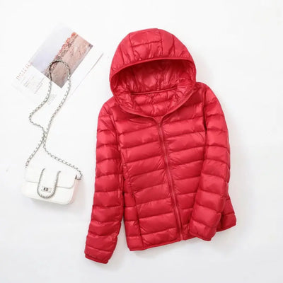Veste doudoune rouge avec capuche sur fond blanc, accompagnée d'un sac à main blanc à chaîne argentée et d'un magazine. Mode hiver femme.