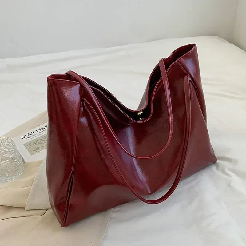 Sac à main en cuir rouge bordeaux élégant sur un lit blanc, avec un livre et un verre à côté. Accessoire mode femme tendance et chic.