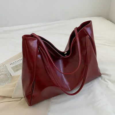 Sac à main en cuir rouge bordeaux élégant sur un lit blanc, avec un livre et un verre à côté. Accessoire mode femme tendance et chic.