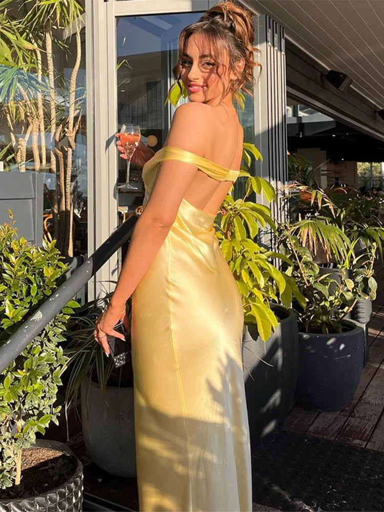 Jeune femme en robe jaune élégante, tenant un verre de vin rosé, posant sur une terrasse ensoleillée entourée de plantes vertes.