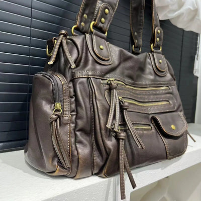Sac à main en cuir marron avec fermetures éclair dorées, poches multiples et détails cloutés, posé sur une surface blanche devant des stores noirs.