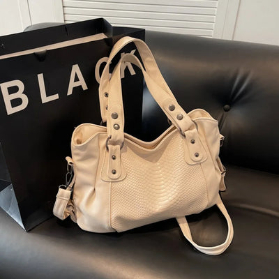 Sac à main beige en cuir texturé avec détails métalliques, posé sur un canapé noir, à côté d'un sac shopping marqué "BLACK". Accessoire mode élégant.
