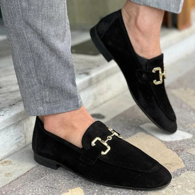 Chaussures mocassins en daim noir pour homme avec boucle dorée, portées avec un pantalon gris. Élégance et style moderne pour tenues formelles.