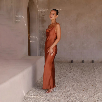 Femme en robe longue satinée marron, posant dans un intérieur minimaliste avec murs en béton. Mode élégante, style chic, tendance robe satin.