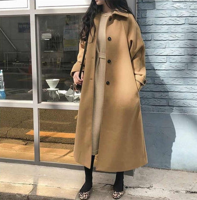 Femme portant un long manteau camel élégant, debout devant une vitrine. Mode automne-hiver, style urbain chic, tendance vêtements femme.