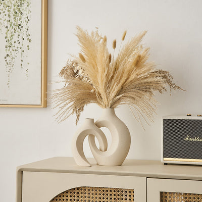 Vase moderne en céramique beige avec pampas séchées sur une commode beige, à côté d'une enceinte vintage et d'un tableau végétal. Décoration intérieure élégante.
