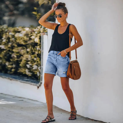 Femme en débardeur noir, short en jean, sandales noires, lunettes de soleil, sac en cuir marron, marchant sur un trottoir ensoleillé.