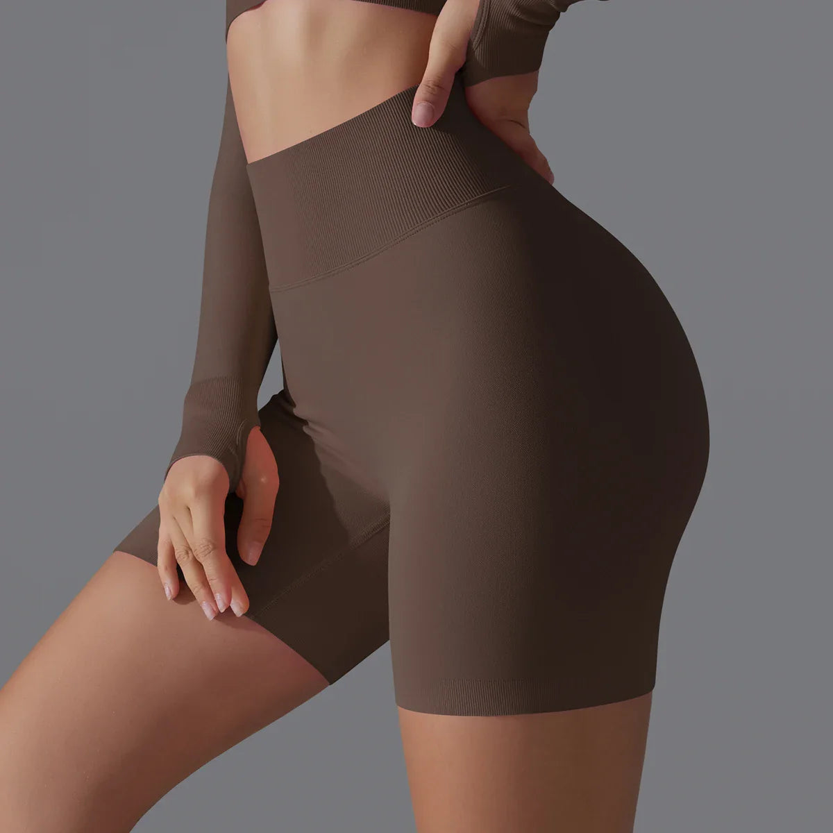 Femme en tenue de sport marron, portant un short taille haute et un haut à manches longues, sur fond gris. Mode fitness, vêtements athlétiques.