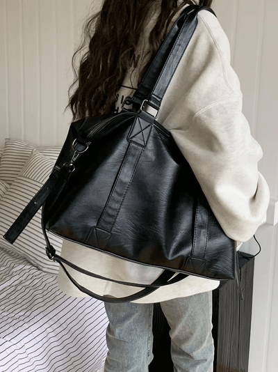 Sac à main noir en cuir porté par une femme en sweat beige, debout près d'un lit à rayures. Mode tendance, accessoire élégant, style décontracté.