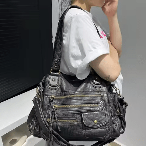 Sac à main en cuir noir avec multiples poches zippées, porté par une femme en t-shirt blanc. Accessoire mode élégant et pratique.