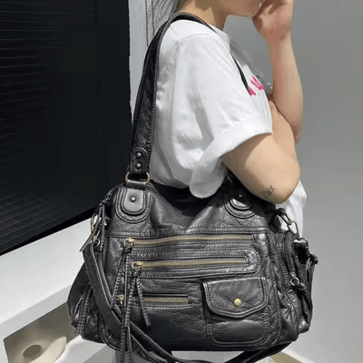 Sac à main en cuir noir avec multiples poches zippées, porté par une femme en t-shirt blanc. Accessoire mode élégant et pratique.