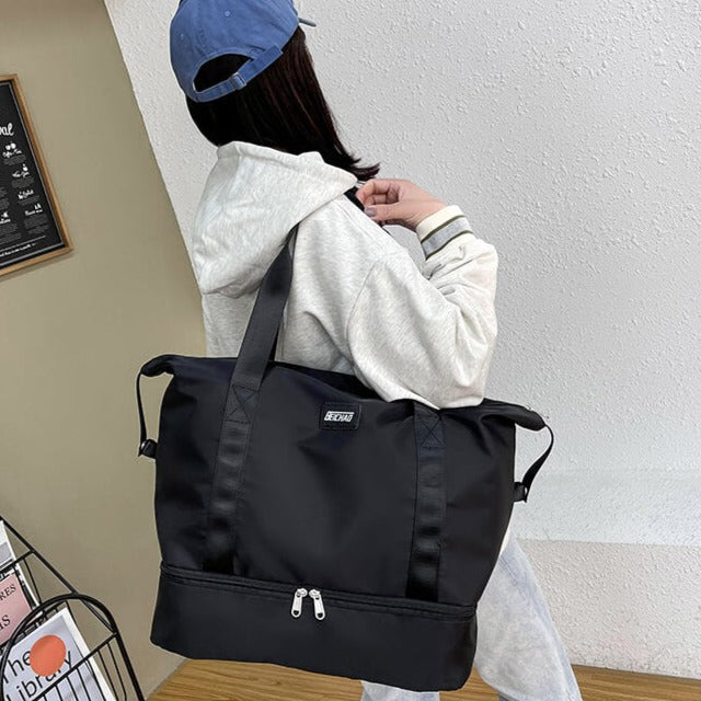 Femme portant un grand sac de sport noir avec fermeture éclair, vêtue d'un sweat à capuche gris et casquette bleue. Mode décontractée, accessoires tendance.