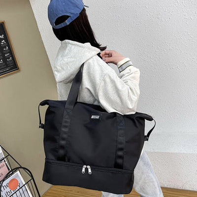 Femme portant un grand sac de sport noir avec fermeture éclair, vêtue d'un sweat à capuche gris et casquette bleue. Mode décontractée, accessoires tendance.