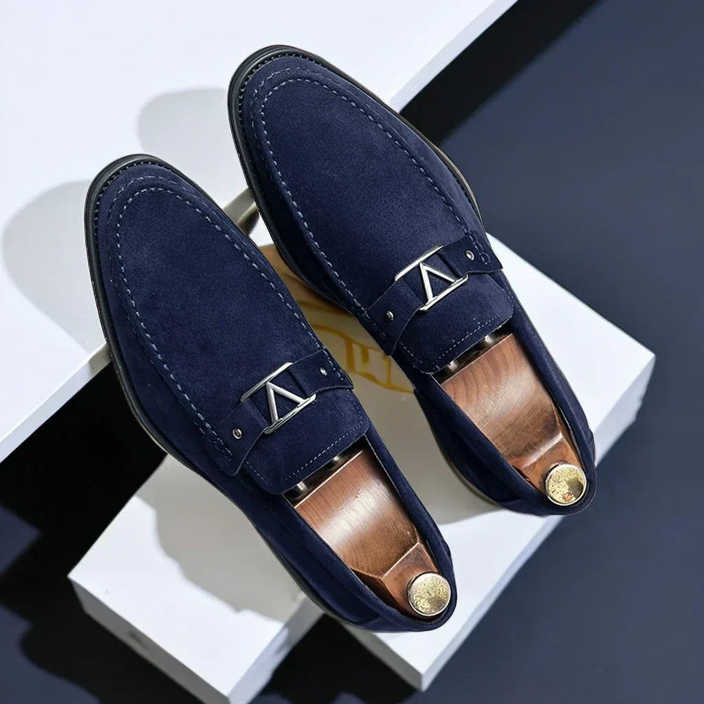 Mocassins en daim bleu marine élégants avec boucle métallique argentée, posés sur fond blanc. Chaussures homme tendance, style chic et moderne.