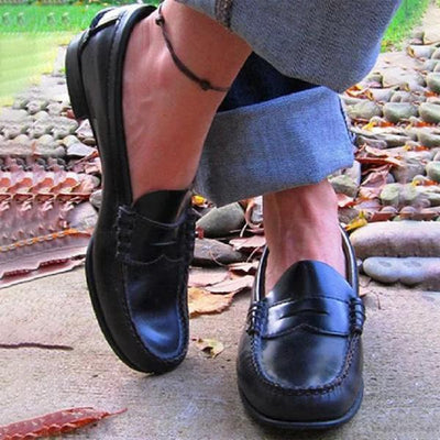 Chaussures mocassins en cuir noir portées avec un jean retroussé, posées sur un chemin de pierres et feuilles d'automne. Mode décontractée élégante.