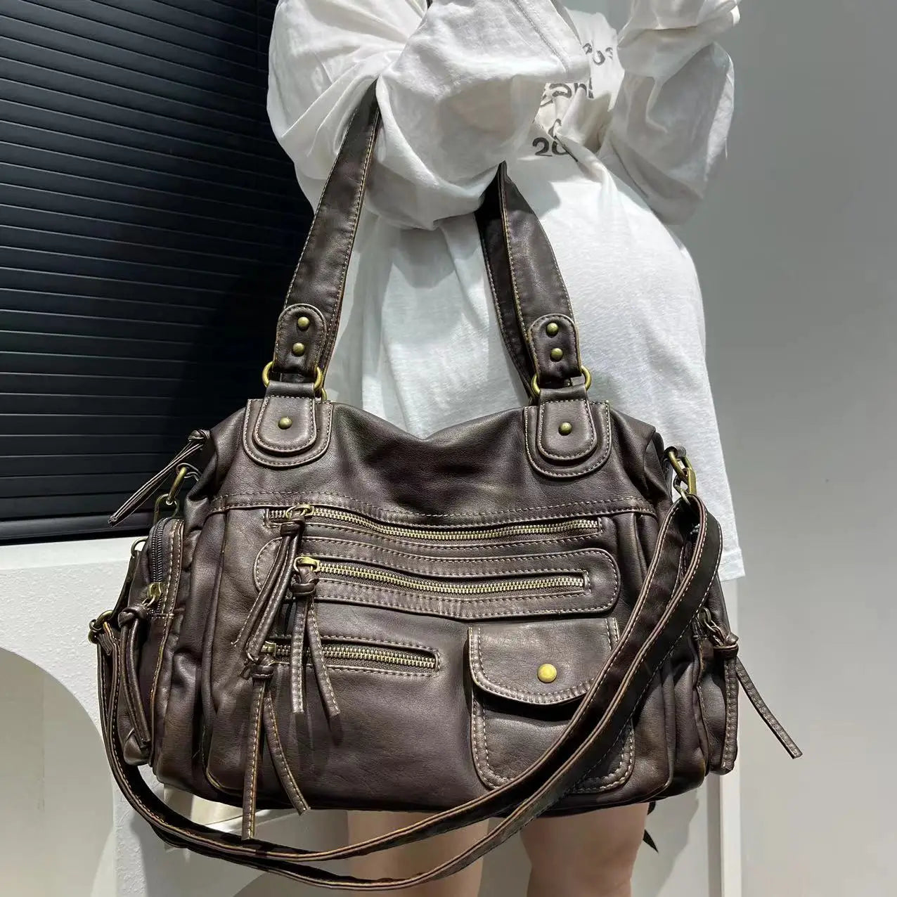 Sac à main en cuir marron avec multiples poches zippées et détails dorés, porté par une personne en tenue blanche. Mode, accessoires, élégance.