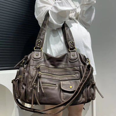 Sac à main en cuir marron avec multiples poches zippées et détails dorés, porté par une personne en tenue blanche. Mode, accessoires, élégance.