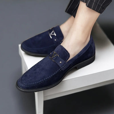 Chaussures mocassins en daim bleu marine pour homme, élégantes et modernes, avec détail métallique, posées sur une table blanche.