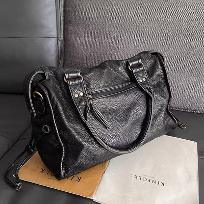 Sac à main en cuir noir élégant sur un canapé, avec des détails métalliques dorés et une poche zippée, posé sur des magazines. Accessoire mode chic.