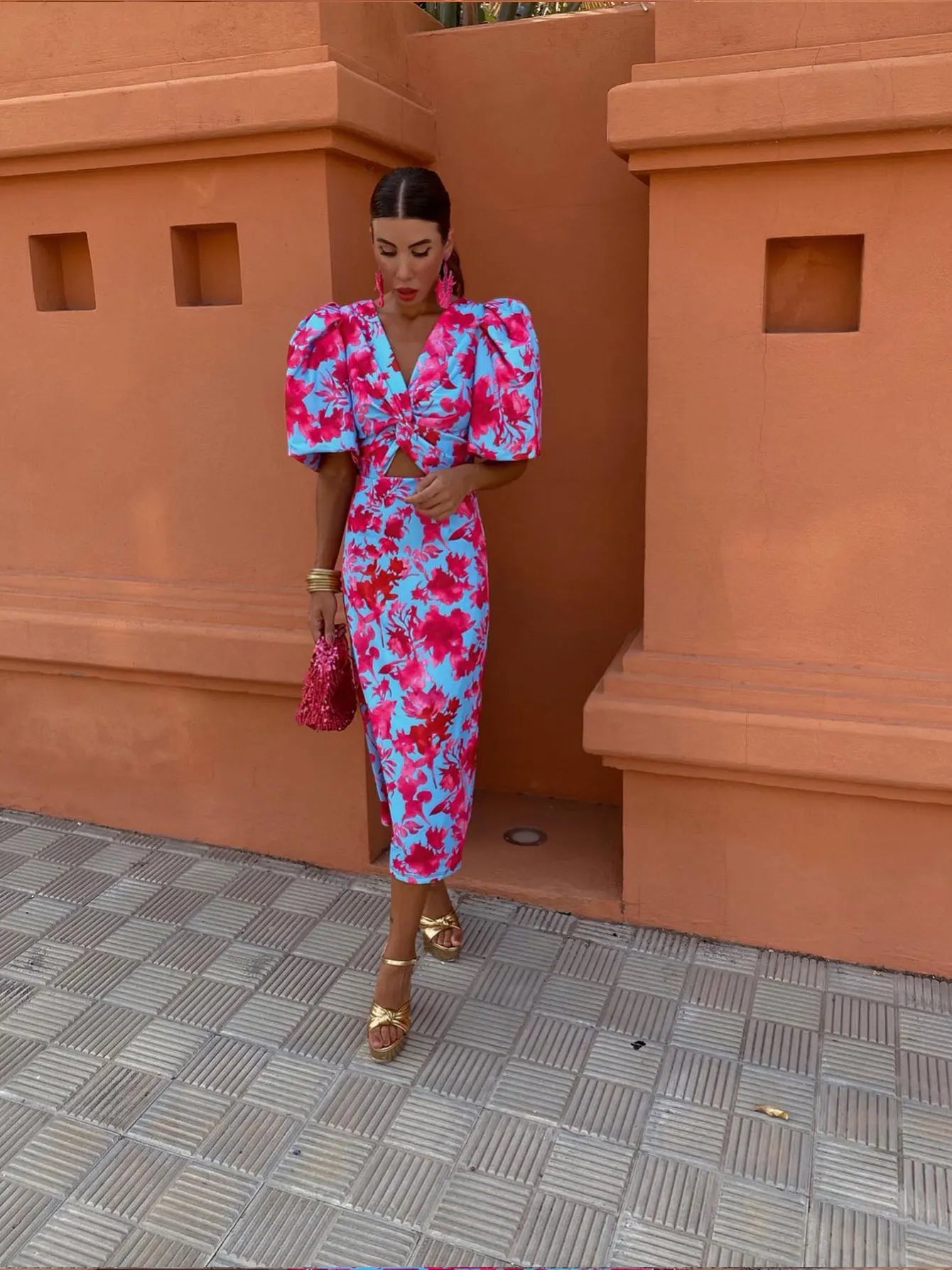 Femme en robe florale rouge et bleue avec manches bouffantes, sac à main rose, sandales dorées, posant devant un mur orange. Mode élégante et colorée.