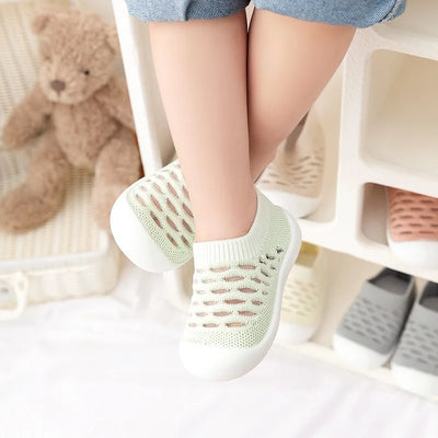Enfant portant des chaussures en maille vertes, semelles blanches, confortables et respirantes, avec un ours en peluche en arrière-plan.