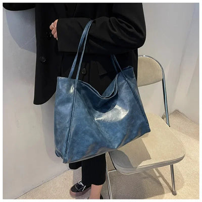 Sac à main bleu en cuir porté par une personne en manteau noir, posé sur une chaise beige. Mode élégante, accessoire tendance pour femme.