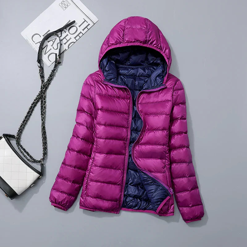 Doudoune violette à capuche, doublure bleue, posée à plat avec un sac à main blanc et noir à côté, sur fond gris. Mode hiver femme.