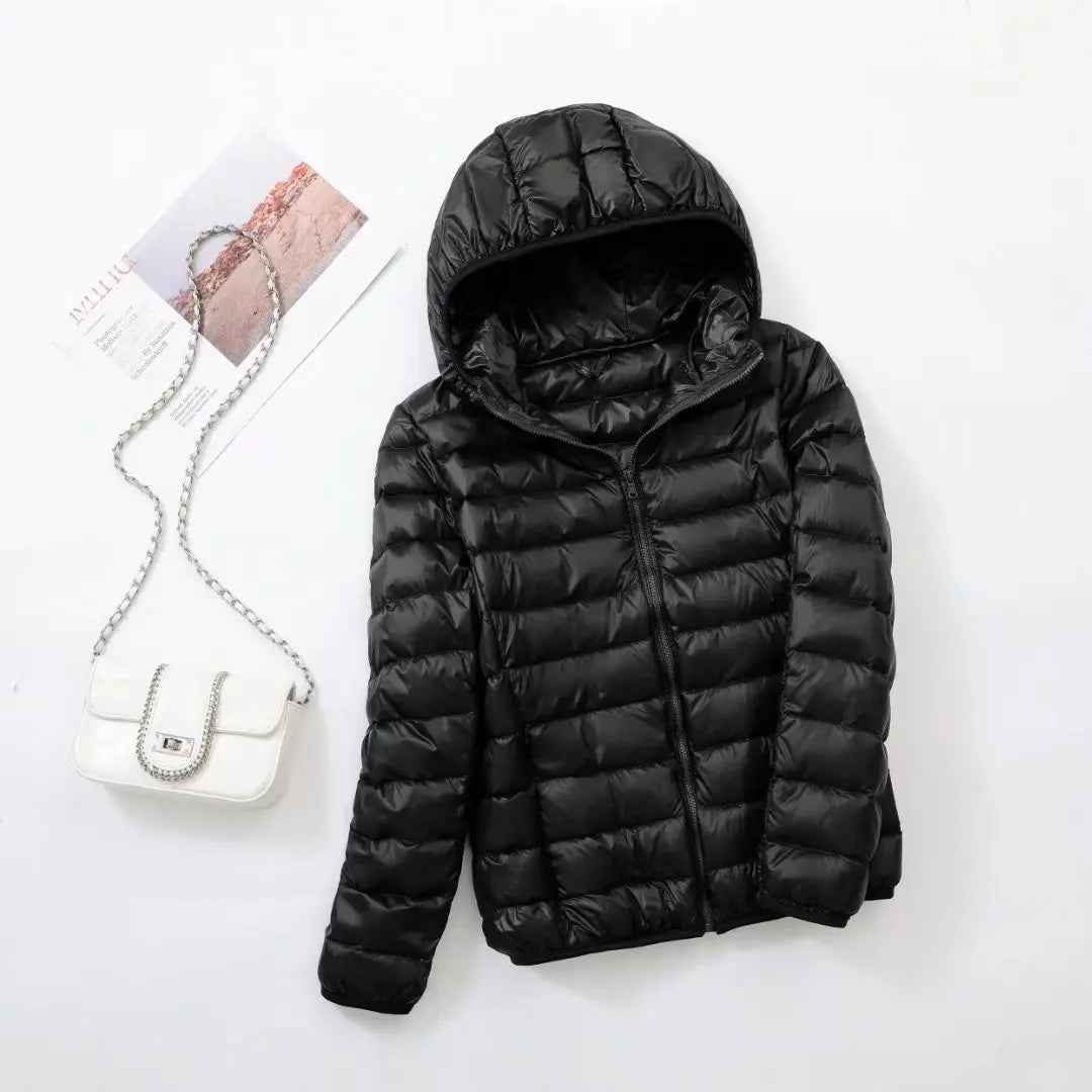 Veste doudoune noire avec capuche sur fond blanc, accompagnée d'un sac à main blanc à chaîne argentée et d'un magazine. Mode hiver élégante.