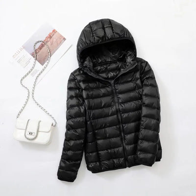 Veste doudoune noire avec capuche sur fond blanc, accompagnée d'un sac à main blanc à chaîne argentée et d'un magazine. Mode hiver élégante.
