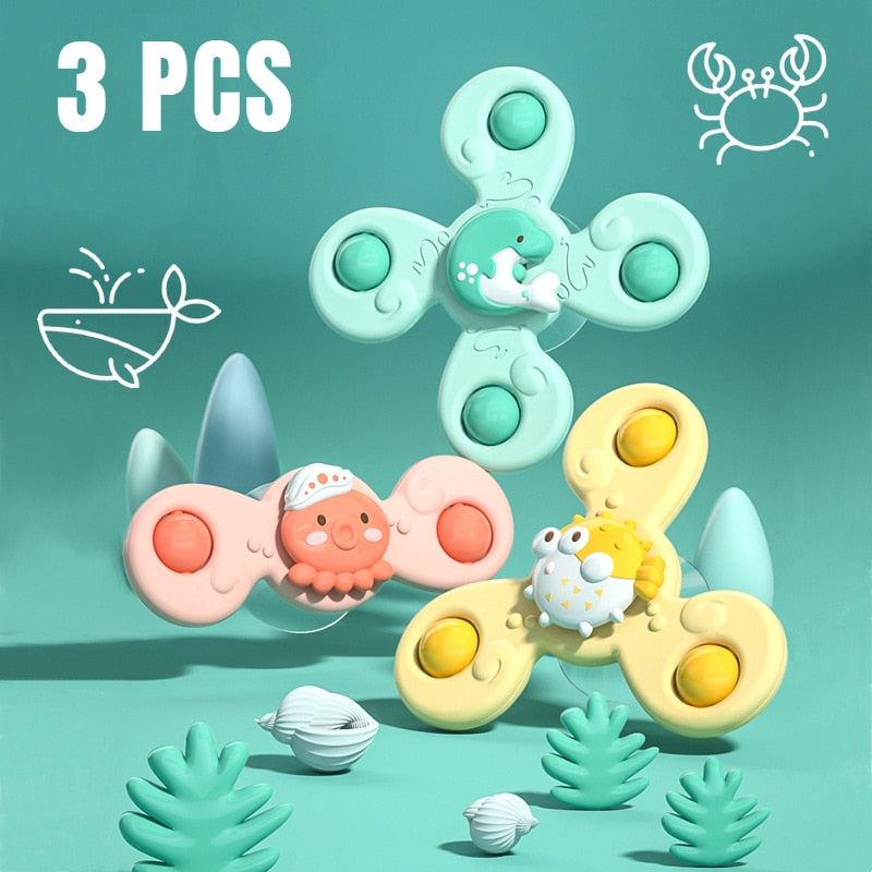 Trois toupies fidget colorées avec motifs animaux marins, dont un crabe et un poisson, sur fond bleu avec coquillages et plantes. Jouet anti-stress.