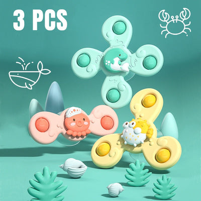 Trois toupies fidget colorées avec motifs animaux marins, dont un crabe et un poisson, sur fond bleu avec coquillages et plantes. Jouet anti-stress.