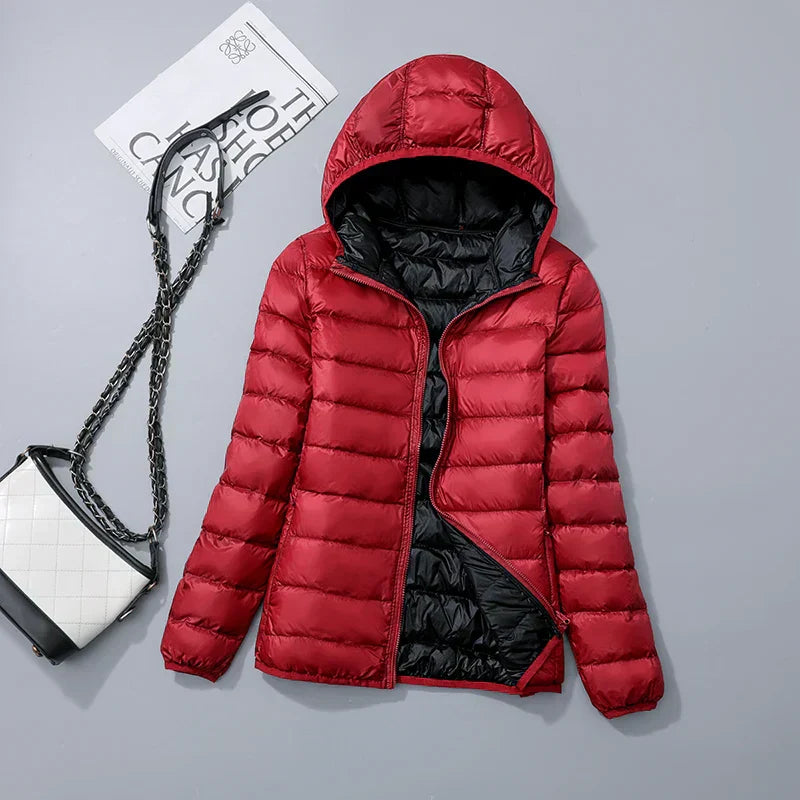 Veste doudoune rouge à capuche, doublure noire, posée à plat avec un sac à main blanc et noir à chaîne, sur fond gris.