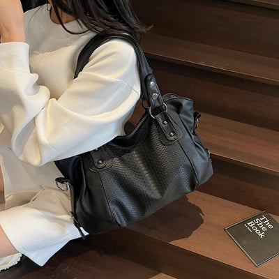 Sac à main noir en cuir texturé, porté par une femme en robe blanche, posé sur des escaliers en bois avec un livre noir à côté. Accessoire mode élégant.